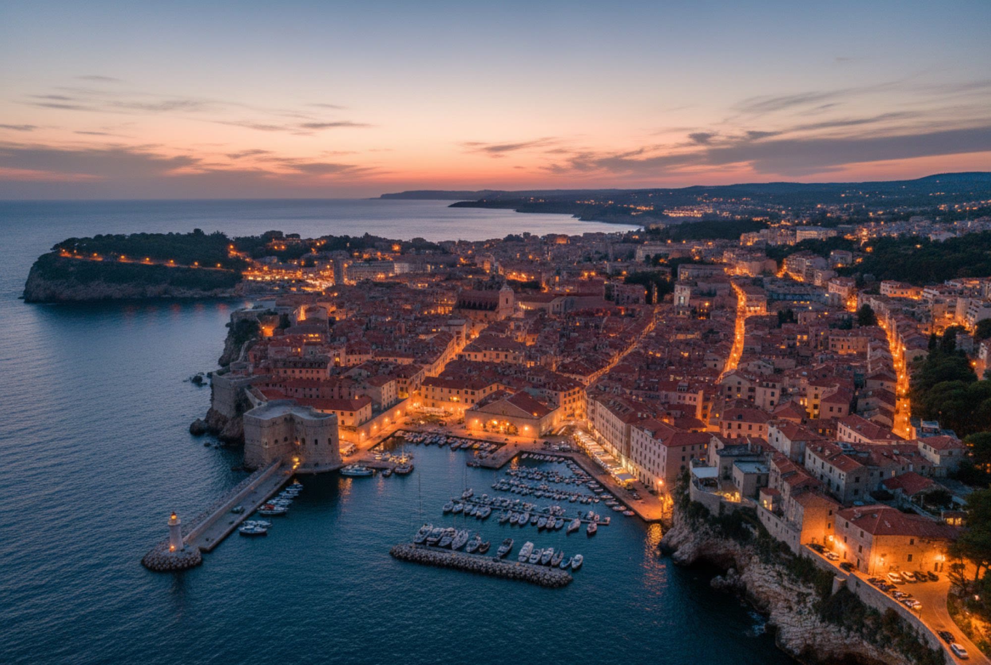 A coastal city at dusk, harbour and rooftops with the lights coming on